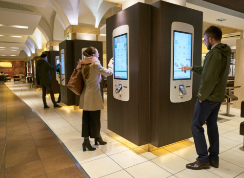 People are using metal touch screen kiosks in a popular fast food establishment.